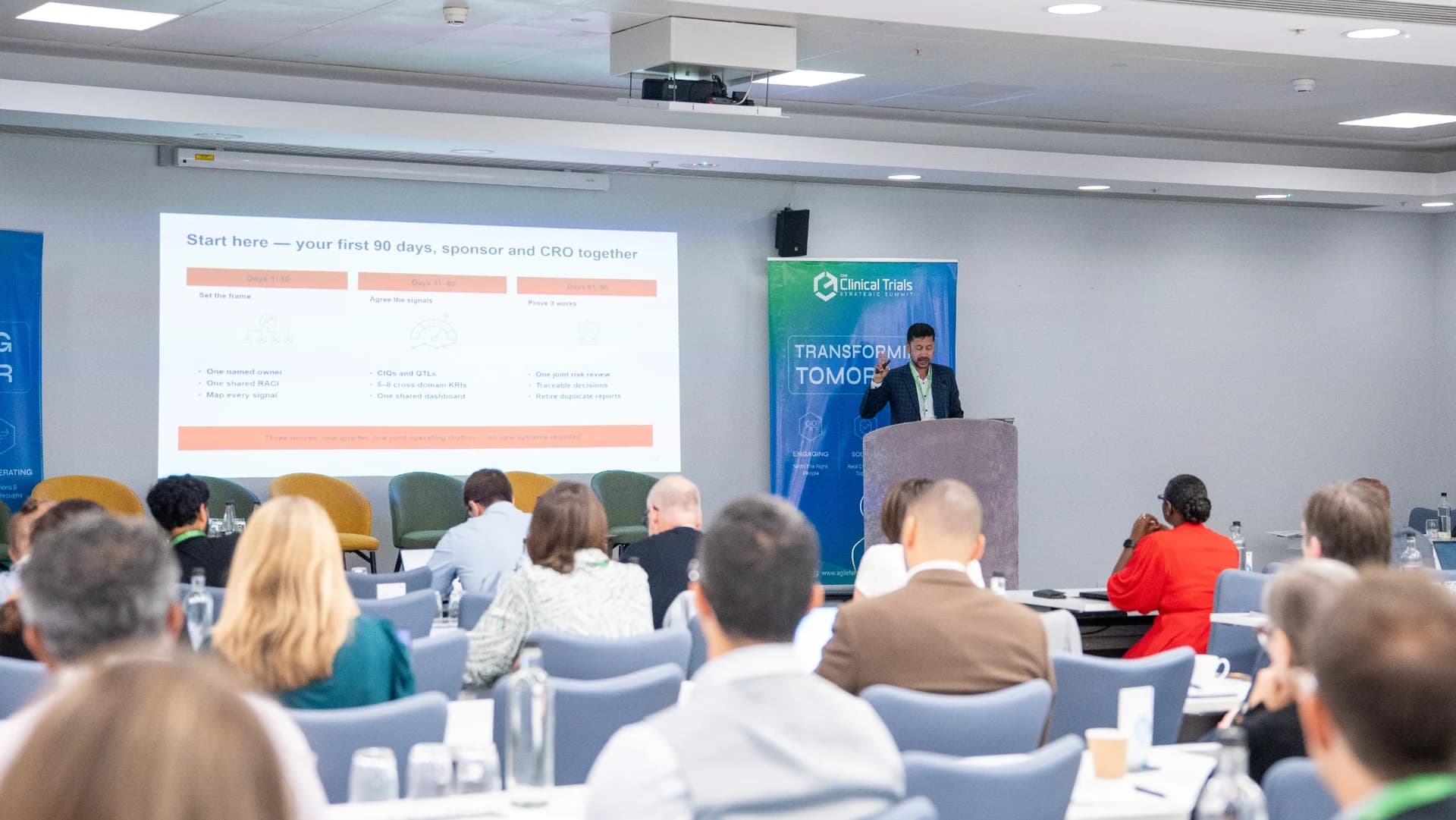 A speaker presenting at the front of a full conference room at the Clinical Trials Strategic Summit